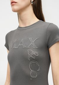 Κοντινό πλάνο ενός ατόμου που φοράει γκρι ριμπ μπλουζάκι με κέντημα "MAX & CO." μπροστά και ασημένια σκουλαρίκια κρίκους.