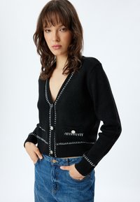 Svart stickad cardigan med dekorativ vit sömnad, frampocket och stora knappar. V-ringad design med långa ärmar.