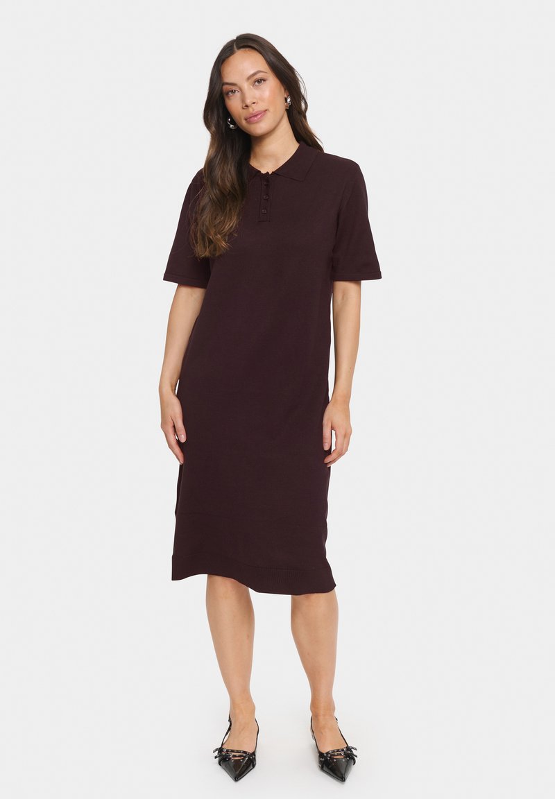 Femme aux longs cheveux bruns portant une robe polo bourgogne foncé à mi-longueur et des chaussures noires à bout pointu, se tenant contre un fond blanc.