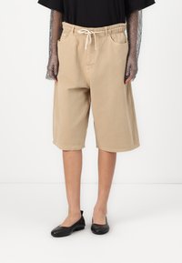 béžové šortky s elastickým páskem, šňůrkou a bočními kapsami. Stylizované s černým topem a černými plochými botami, na bílém pozadí.