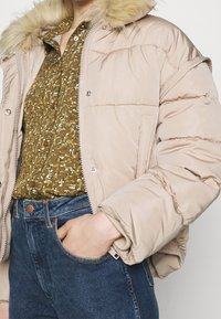Beige dunjakke med hætte foret med pels, lynlås fortil og ribbede manchetter, sat sammen med en grøn mønstret bluse og blå højtaljede jeans.