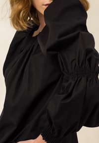 Frau in einer schwarzen Bluse mit voluminösen Puffärmeln und gerafften Manschetten, die teilweise ihr Gesicht mit ihrem Arm verdeckt.