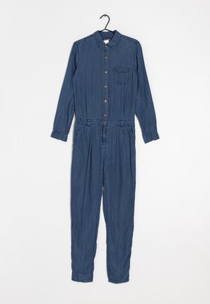 Salopette en denim bleu à manches longues avec des boutons à l'avant, une poche poitrine et des poches latérales, accrochée à un porte-manteau noir sur fond blanc.