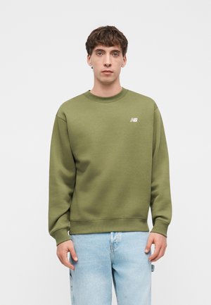 Joven con pelo corto y marrón, vistiendo una sudadera verde oliva de New Balance y vaqueros azul claro, posando contra un fondo liso.