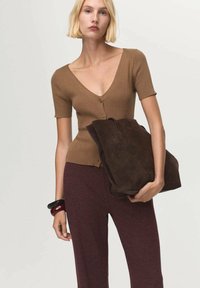 Brown ribbed short-sleeve cardigan with a V-neck, button front, paired with burgundy trousers. A dark brown suede tote bag is held in one hand.