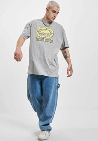 Grijze oversized t-shirt met een gele grafische print, gedragen met losse blauwe jeans en witte sneakers. Korte mouwen en een ontspannen pasvorm.