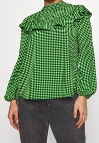 Green checkered blouse with ruffle accents, high neckline, and long sleeves. Fabric appears lightweight and textured with gathered cuffs.