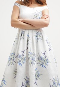 Robe blanche à  épaules dénudées avec un imprimé floral en bleu et vert, tissu texturé, jupe ample et taille froncée.