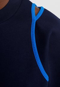 Gros plan sur un tissu bleu foncé avec une couture zigzag bleu vif soulignant une découpe à l'épaule sur un vêtement.