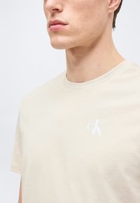 Νεαρός άνδρας που φοράει ένα απλό μπεζ μπλουζάκι με λευκό λογότυπο "cK" στο αριστερό στήθος, σε ανοιχτό φόντο.