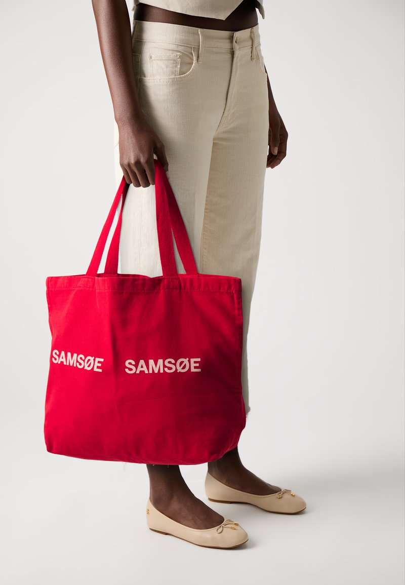 Red canvas tote bag with "SAMSØE" printed in white. Features sturdy shoulder straps and a simple, rectangular design.