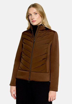Femme aux cheveux blonds portant une veste marron matelassée et lisse avec une fermeture éclair à l'avant et un pantalon noir sur un fond clair.