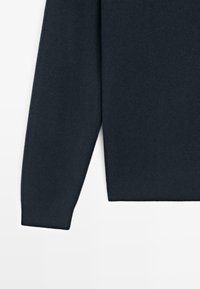 Tmavě modrý svetr s hladkou texturou, žebrovanými manžetami a lemem. Má klasický kulatý výstřih a dlouhé rukávy. Materiál se zdá být měkký a pletený.