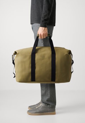 Una persona con jeans grigi e sneakers tiene in mano una grande borsa a tracolla verde oliva con cinghie nere e il logo RAINS.