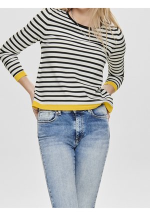 Femme portant un t-shirt à manches longues rayé noir et blanc avec une bordure jaune et un jean bleu clair délavé, les mains dans les poches, sur un fond uni.