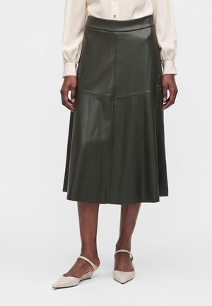 Femme portant une jupe en cuir vert foncé longueur genou, un chemisier blanc à manches longues, et des talons bas beiges avec des brides à la cheville.