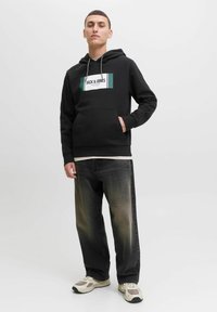 Schwarzer Kapuzenpullover mit Fronttasche und Kordeln, mit einem weißen und türkisfarbenen grafischen Logo. Kombiniert mit ausgebleichten schwarzen Jeans und beigen Sneakers.