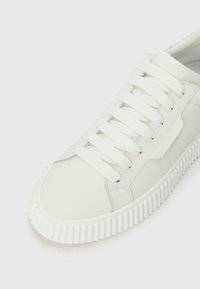 Sneaker alb cu talpă groasă cu nervuri, șireturi albe și branding subtil pe lateral, pe un fundal luminos și simplu.