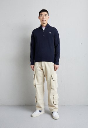 Navy zip-up sweatshirt with a small logo, paired with cream cargo pants featuring multiple pockets and white sneakers.