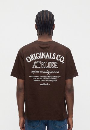 Person mit geflochtenen Haaren, die ein braunes T-Shirt mit weißer Aufschrift Originals Co. Atelier und blaue Jeans trägt, mit dem Rücken zur Kamera.