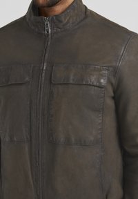 Braune Lederjacke mit Stehkragen, frontalem Reißverschluss und zwei Brusttaschen. Die Struktur sieht leicht abgetragen aus, mit sichtbaren Nähdetails.