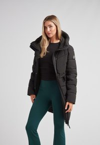 Kvinne iført en svart puffjakke med hette, svart topp og teal bukser, som står mot en ensfarget lys grå bakgrunn.