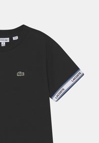 Černé bavlněné tričko s zeleným logem krokodýla na hrudi a modro-bílým rámečkem na rukávech. Klasický design s kulatým výstřihem.