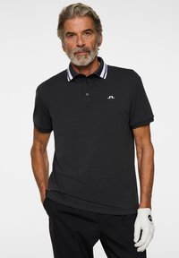 Musta poolopaita, jossa valkoraidallinen kaulus, lyhyet hihat ja rintalogo. Yhdistetty mustiin housuihin ja valkoiseen golf-käsineeseen.