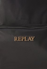 Zaino in tessuto nero con un logo "REPLAY" dorato in evidenza. Dettaglio della tasca con zip lungo il bordo superiore. Superficie liscia e texture.