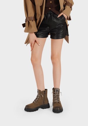 Stivaletti alla caviglia in suede marrone con lacci argentati, tacco testurizzato e suola in gomma. Indossati con pantaloni shorts in pelle nera su uno sfondo neutro.