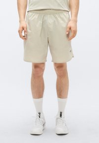 Bež kratke hlače z elastičnim pasom, z gladko teksturo in majhnim logotipom na strani, kombinirane z belimi nogavicami in supergami.