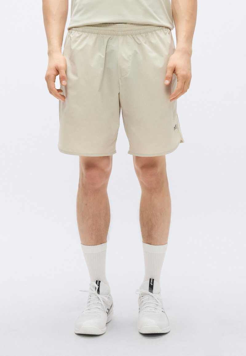 Bež kratke hlače z elastičnim pasom, z gladko teksturo in majhnim logotipom na strani, kombinirane z belimi nogavicami in supergami.