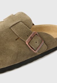 Gros plan d’une sandale en daim vert olive avec boucle en cuivre ajustable et semelle noire sur fond gris clair.