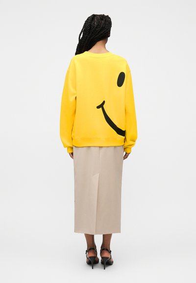 Personne avec de longues tresses portant un sweat-shirt jaune arborant un grand smiley noir au dos et une jupe midi beige avec des talons noirs.