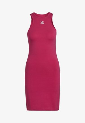 Kobieta ubrana w bezrękawnikową, jasnoróżową sukienkę Adidas oraz białe tenisówki, stojąca z rękami na biodrach na jednolitym tle.