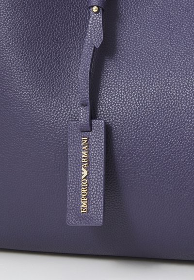 Purple leather handbag featuring a gold emblem and tag that reads "Emporio Armani." Close-up view highlights texture and detail.