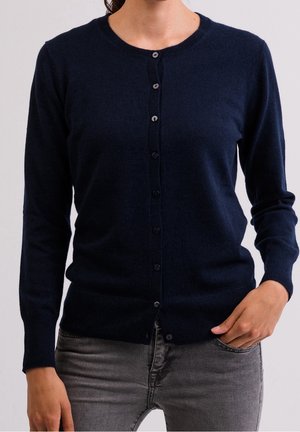 Femme portant un cardigan bleu marine boutonné et un jean gris, une main dans la poche et l'autre détendue le long du corps.