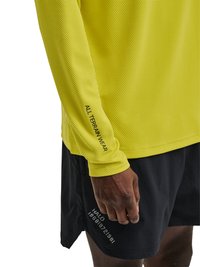 Nahaufnahme einer Person, die ein gelbes, strukturiertes Langarmshirt mit dem Schriftzug "ALL-TERRAIN WEAR" und schwarze Shorts mit reflektierendem "HALO" und Zahlen trägt.