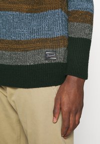 Pull en maille avec des rayures horizontales en bleu, marron, gris et vert. Comprend un patch de marque près de l'ourlet. Poignets et bas côtelés.