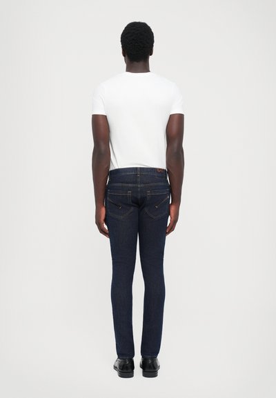 Jeans de mezclilla azul oscuro con un corte ajustado, costuras detalladas y dos bolsillos traseros. Combinados con una camisa blanca básica y zapatos negros.