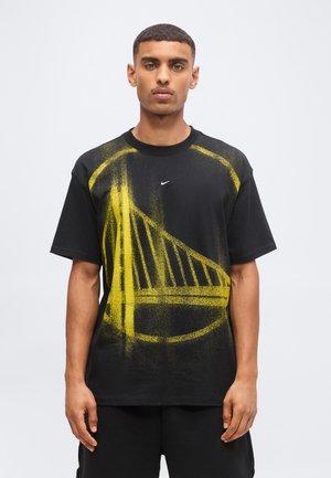 Мъж, носещ черна тениска Nike с голяма жълта графика, наподобяваща мост отпред, стои пред обикновен светъл фон.