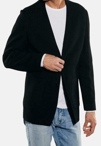 Czarna teksturowana marynarka z klapami z wycięciem, zapinana na pojedynczy guzik, z dwoma przednimi kieszeniami, noszona na białej koszuli i niebieskich dżinsach.