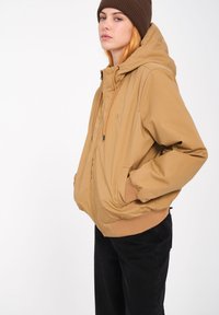 TAN hoodie met een rits, elastische manchetten en twee voorkeuzezakken. Gecombineerd met zwarte broeken en een donkergroene beanie. Eenvoudig ontwerp.