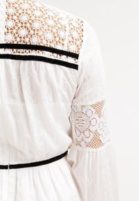 Vestido blanco con detalle de encaje en la parte posterior, acentos de terciopelo negro en los hombros y la cintura, tejido texturizado, mangas largas y patrones de encaje floral.