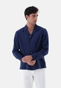 Camicia da uomo a maniche lunghe di colore navy in tessuto leggero, con chiusura a bottoni, colletto a punte e una sottile trama a righe. Sotto si indossano pantaloni bianchi.