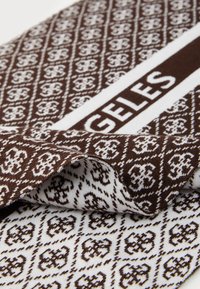 Brun og hvit mønstret stoff med intrikate design. Har et iøynefallende hvitt bånd trykket med teksten "GELES." Myk tekstur, brettet hjørne.