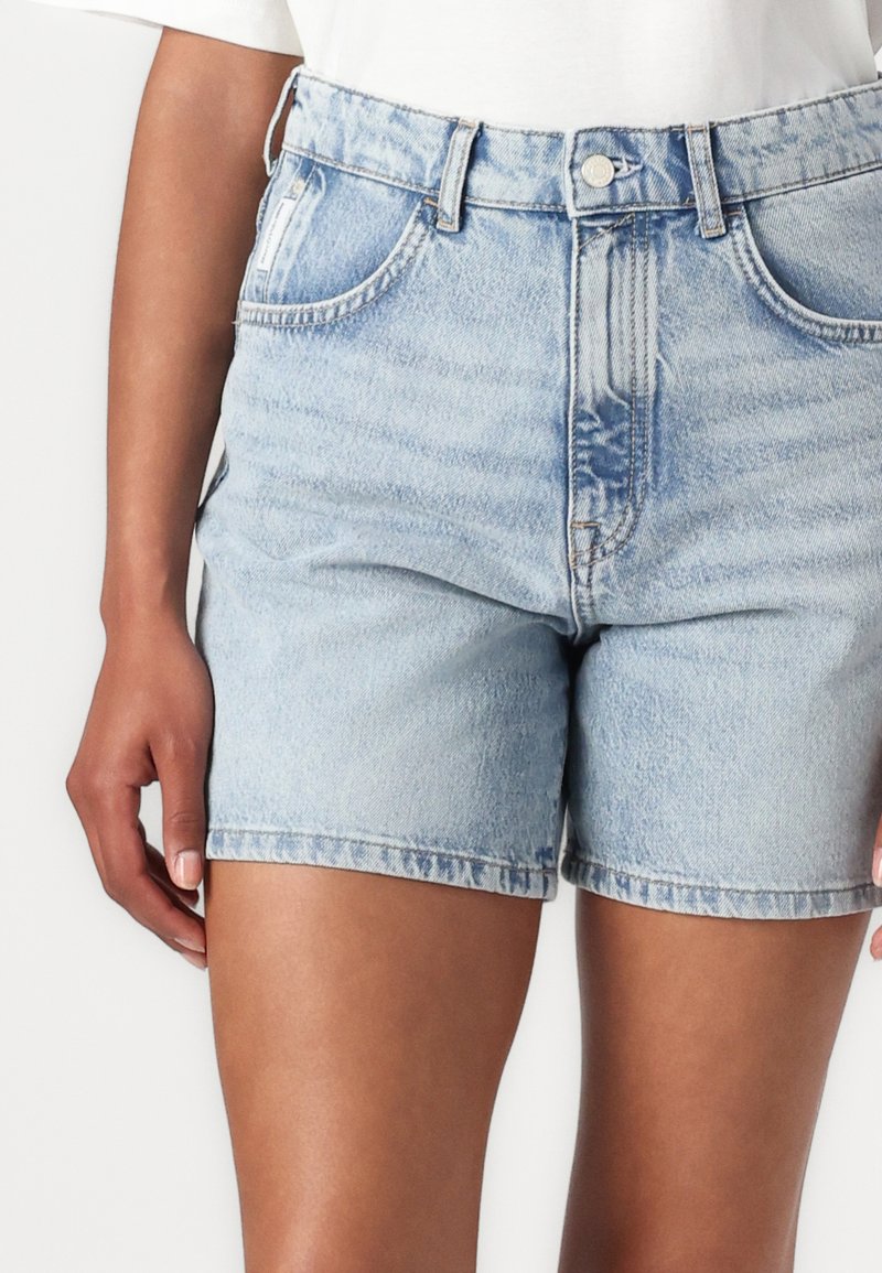 Hellblaue Jeansshorts mit hoher Taille, fünf Taschen, umgeschlagenem Saum und leichter Ausbleichung. Kombiniert mit einem weißen Oberteil.