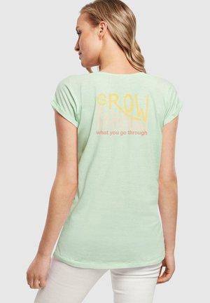 Hellgrünes T-Shirt mit aufgerollten Ärmeln. Auf dem Rücken steht in fetter Schrift: "GROW THROUGH" in Gelb und Pink, mit "what you go through" darunter.