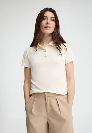 Kvinde med langt brunt hår iført en cremefarvet kortærmet polo og beige højttaljede bukser, stående mod en hvid baggrund.