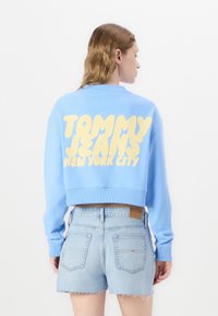 Zils apgrieztais krekls ar dzelteno tekstu "TOMMY JEANS NEW YORK CITY." Iekļauj ribotas aproces un malējās daļas. Saskaņots ar gaiši ziliem džinsu šortiem.